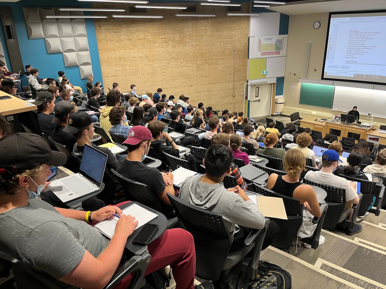 A tiered lecture hall is filled with students sitting in chairs, looking at the lecture screen.