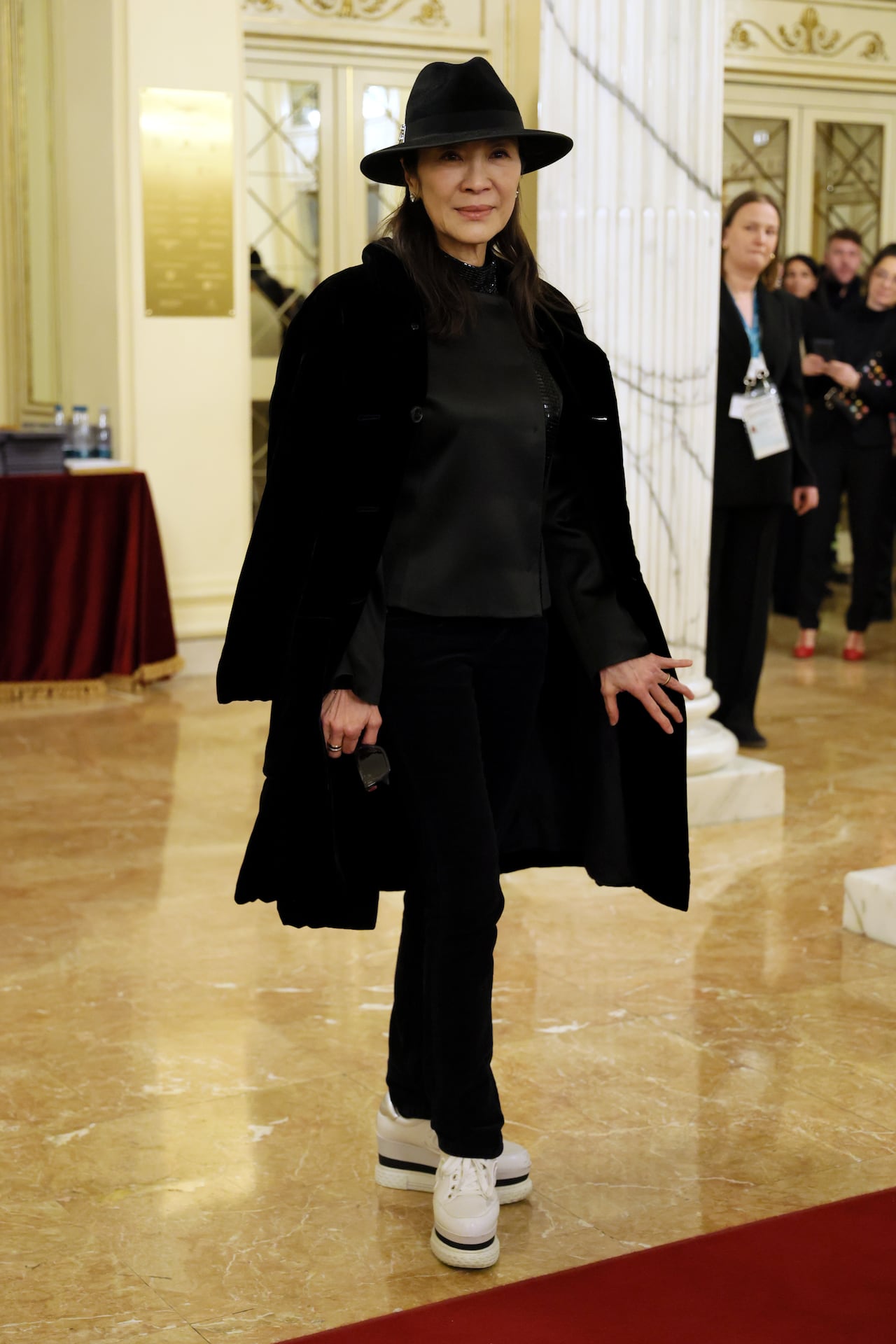 A woman poses for a photo wearing a black hat, jacket and pants with white sneakers. 
