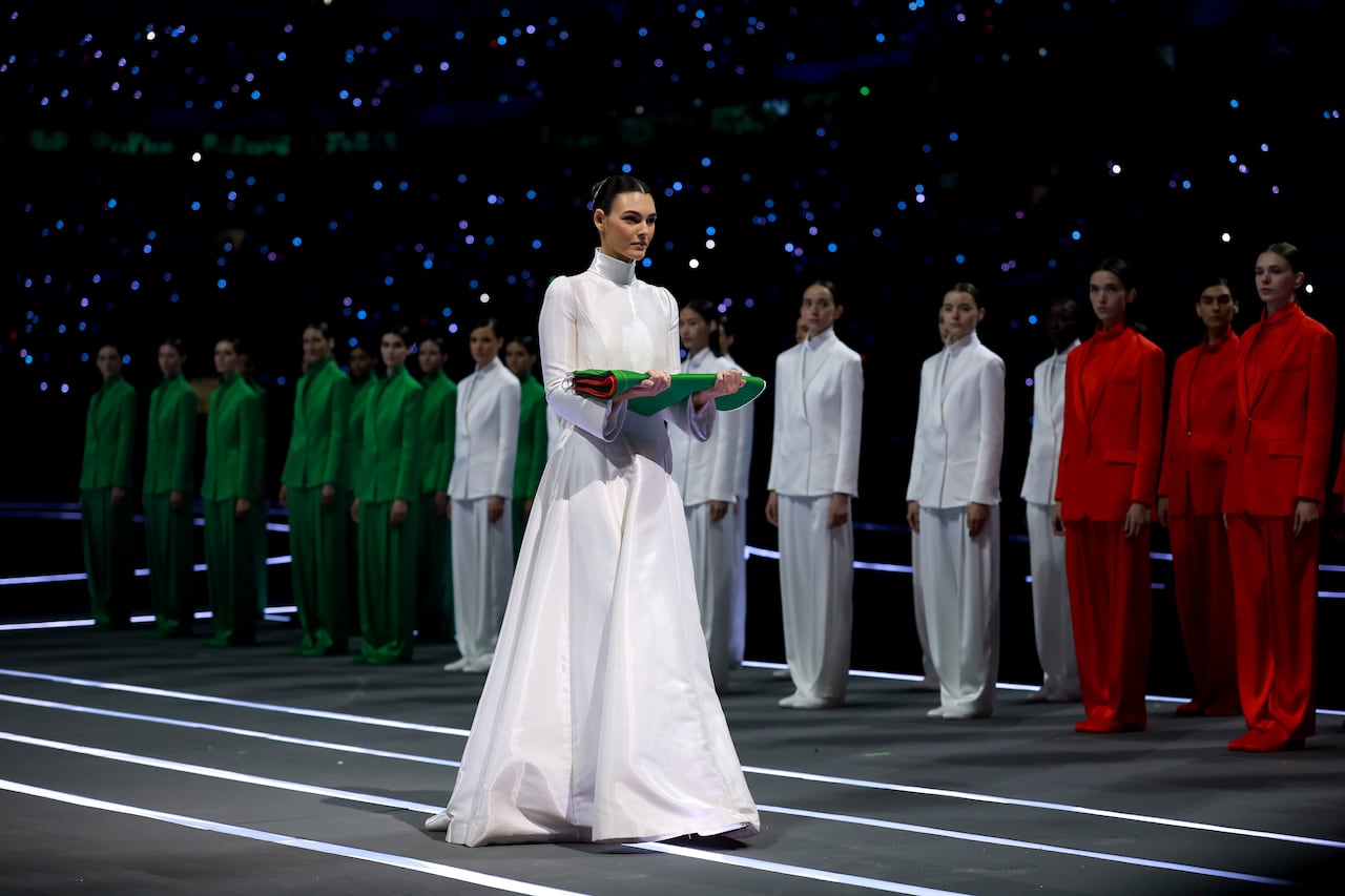 A woman wearing a white gown carries a folded flag and performers wearing red, white or green suits are on stage behind her.