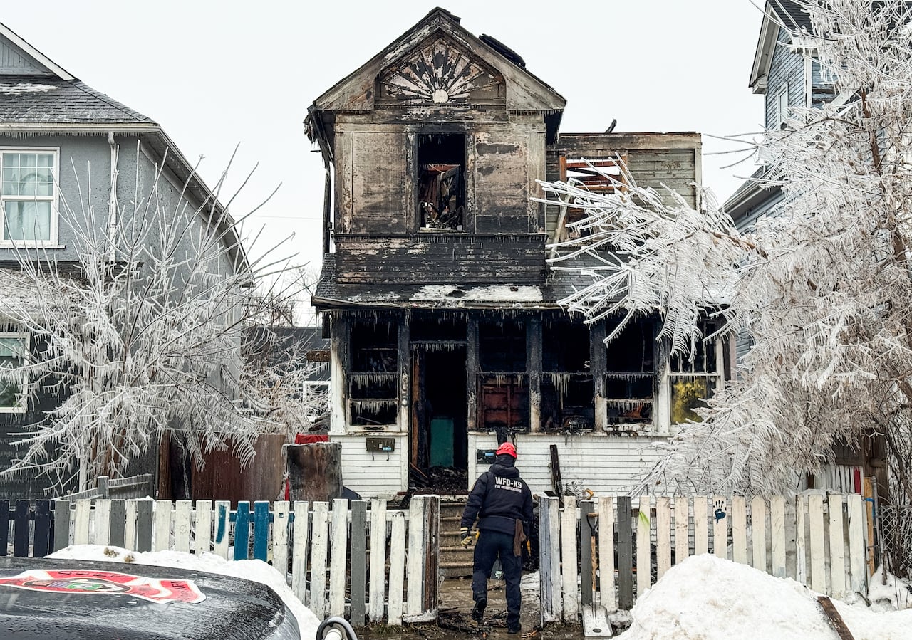A fire official wearing a navy blue winter suit and a red helmet stands outside a charred white house