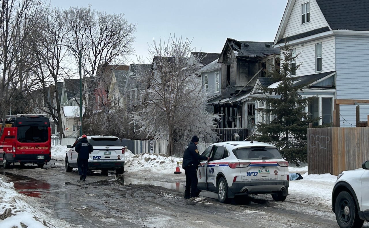 Several vehicles, including a small red fire truck are seen parked on a residential street next to a charred and burned down house