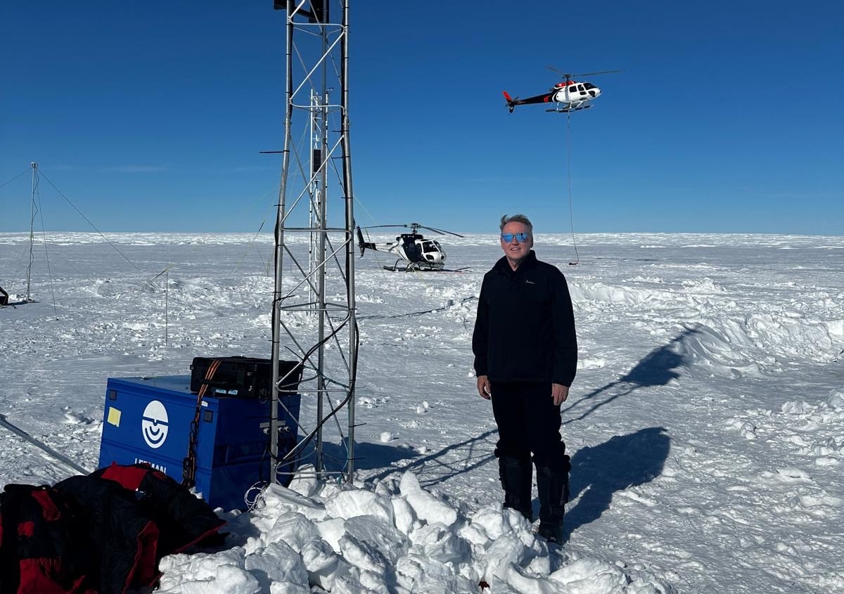 A man stands on a glacier next to a metal tower, a helicopter flies behind him.