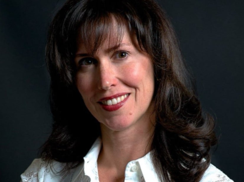 A white woman with long brown hair and a white blouse smiles at the camera.