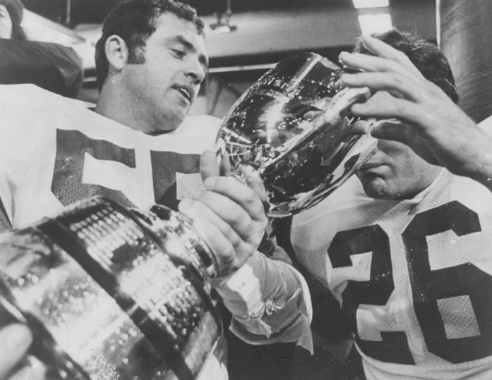 A black-and-white image of two men wearing football jerseys. One of them is holding a trophy while another man is lowering his face toward the trophy. 