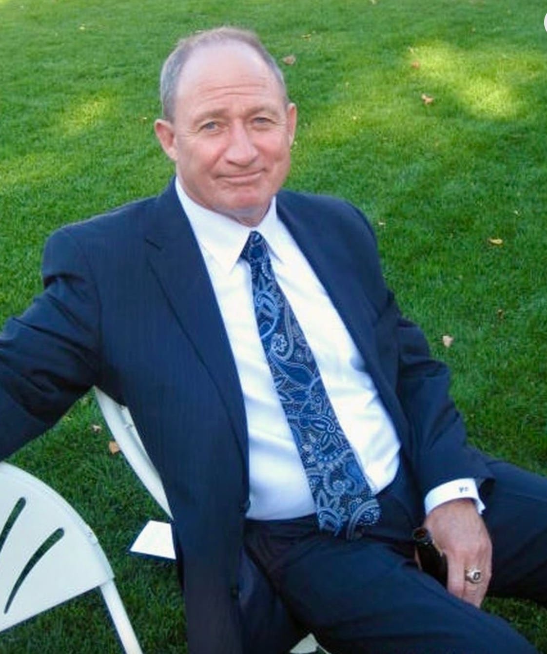 A smiling man wearing a gold ring and a blue suit sits on an outdoor chair.