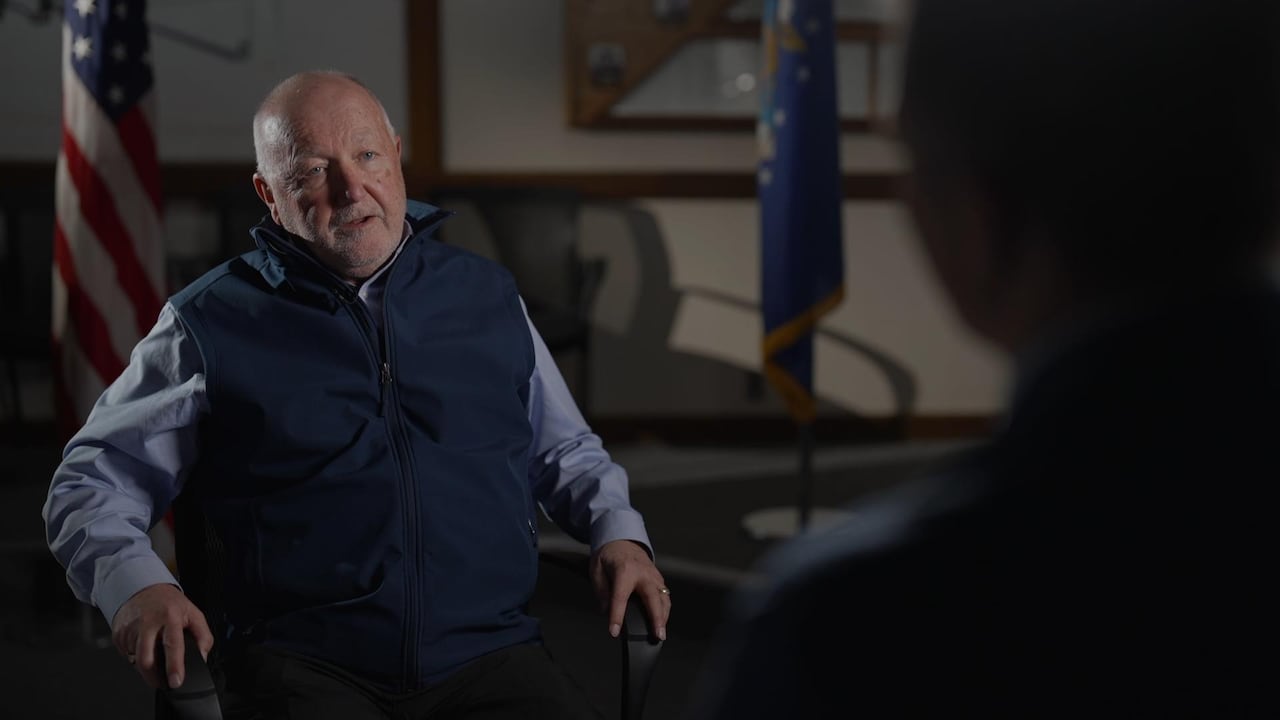 A bald man in a blue collared shirt with a navy fleece sits in a chair in front of an American flag.