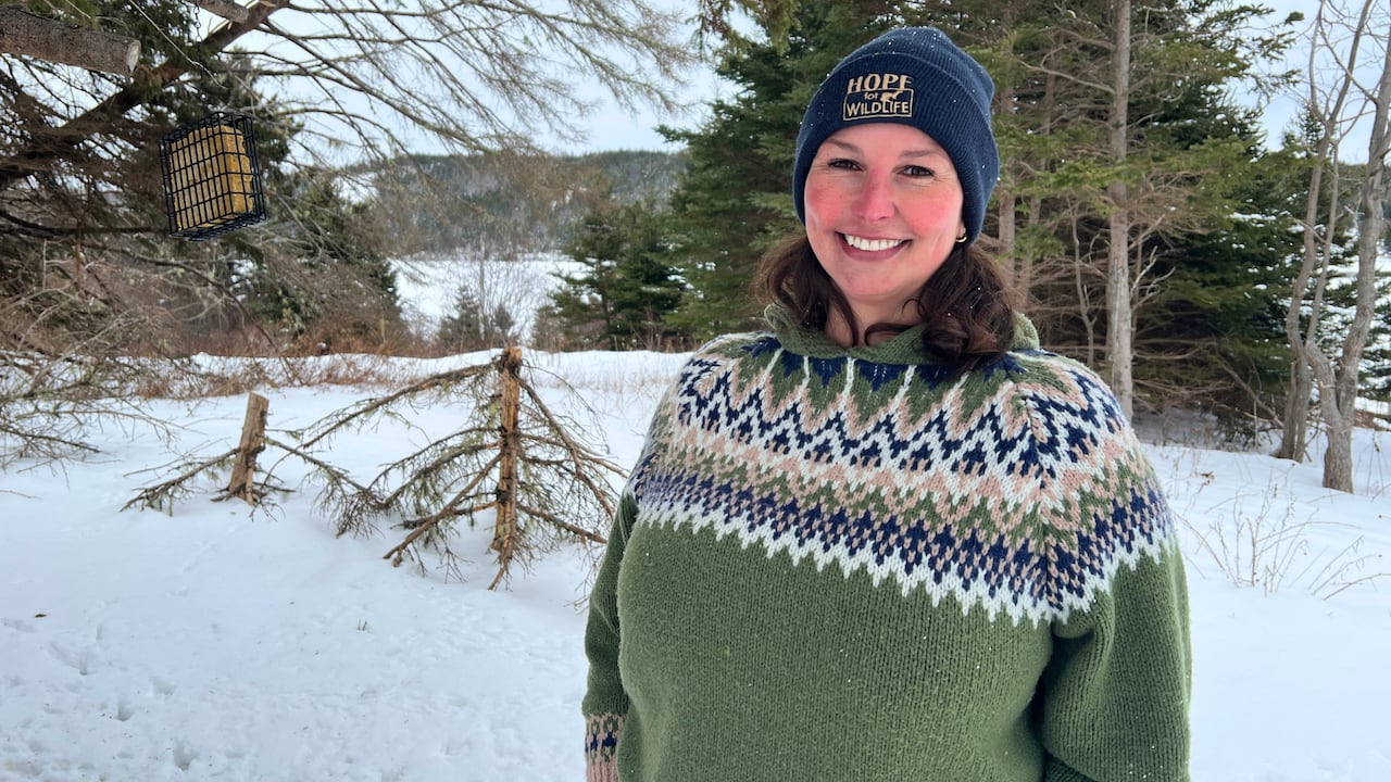 A woman in a green sweater and a hat smiles at the camera. Behind her, snow and trees. 