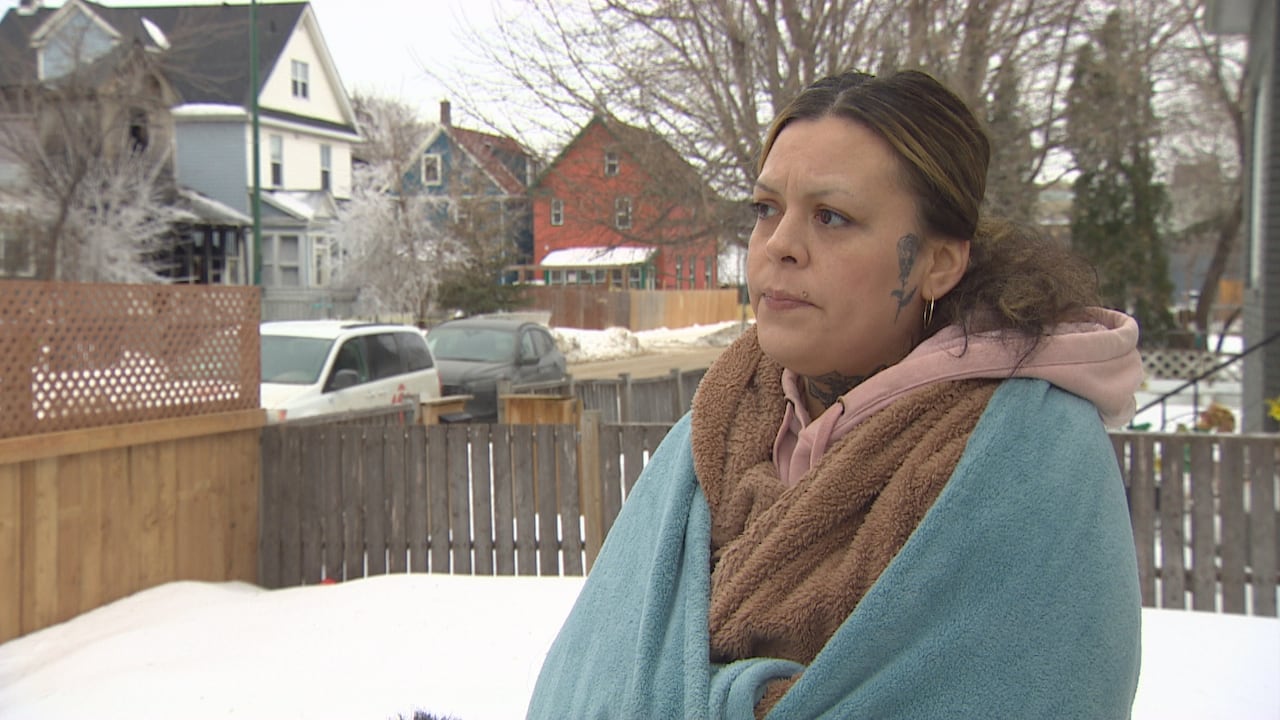 woman with hair in a bun wears several layers of fuzzy blankets while standing outside in a snowy yard on a residential street