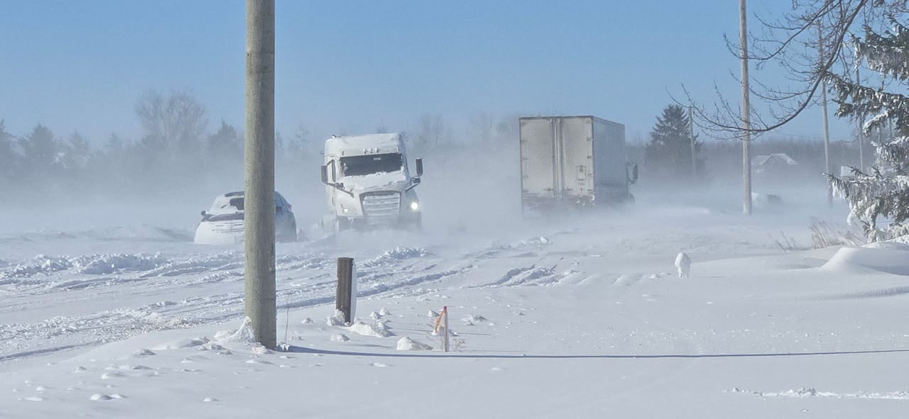 Two two trucks driving on very snowy road, car parked nearby