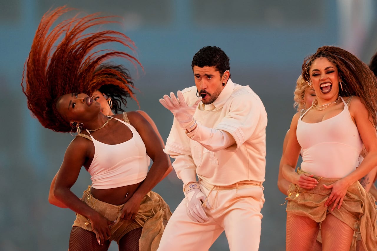 a man in a white outfit surrounded by female backup dancers in matching white tank tops and tan skirts dance on stage during a music performance