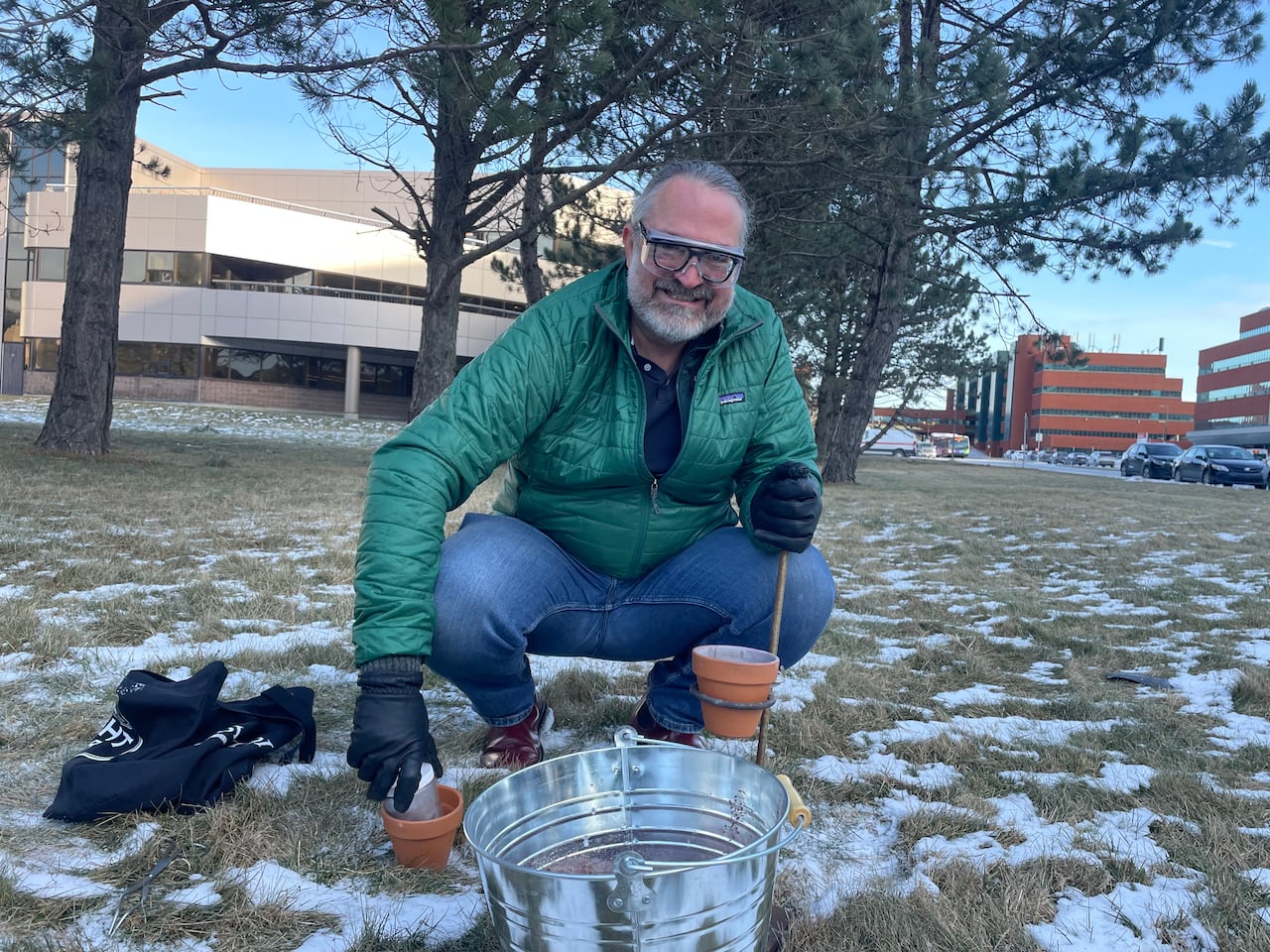 Man in great winter coat crouching down in front of a metal bucket and two small clay pots.
