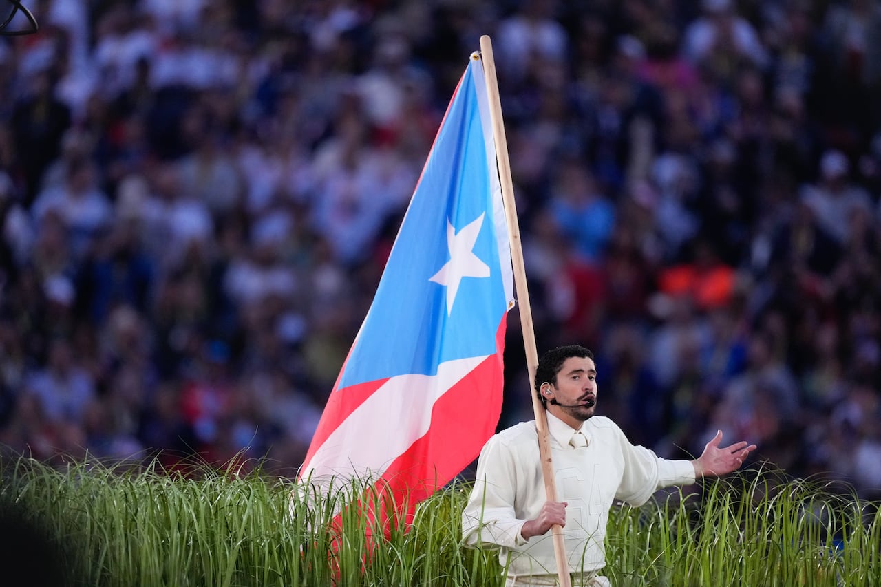 a man in a white outfit sings and carries a massive puerto rican flag that trails behind him as he walks through a set designed with fake grasses