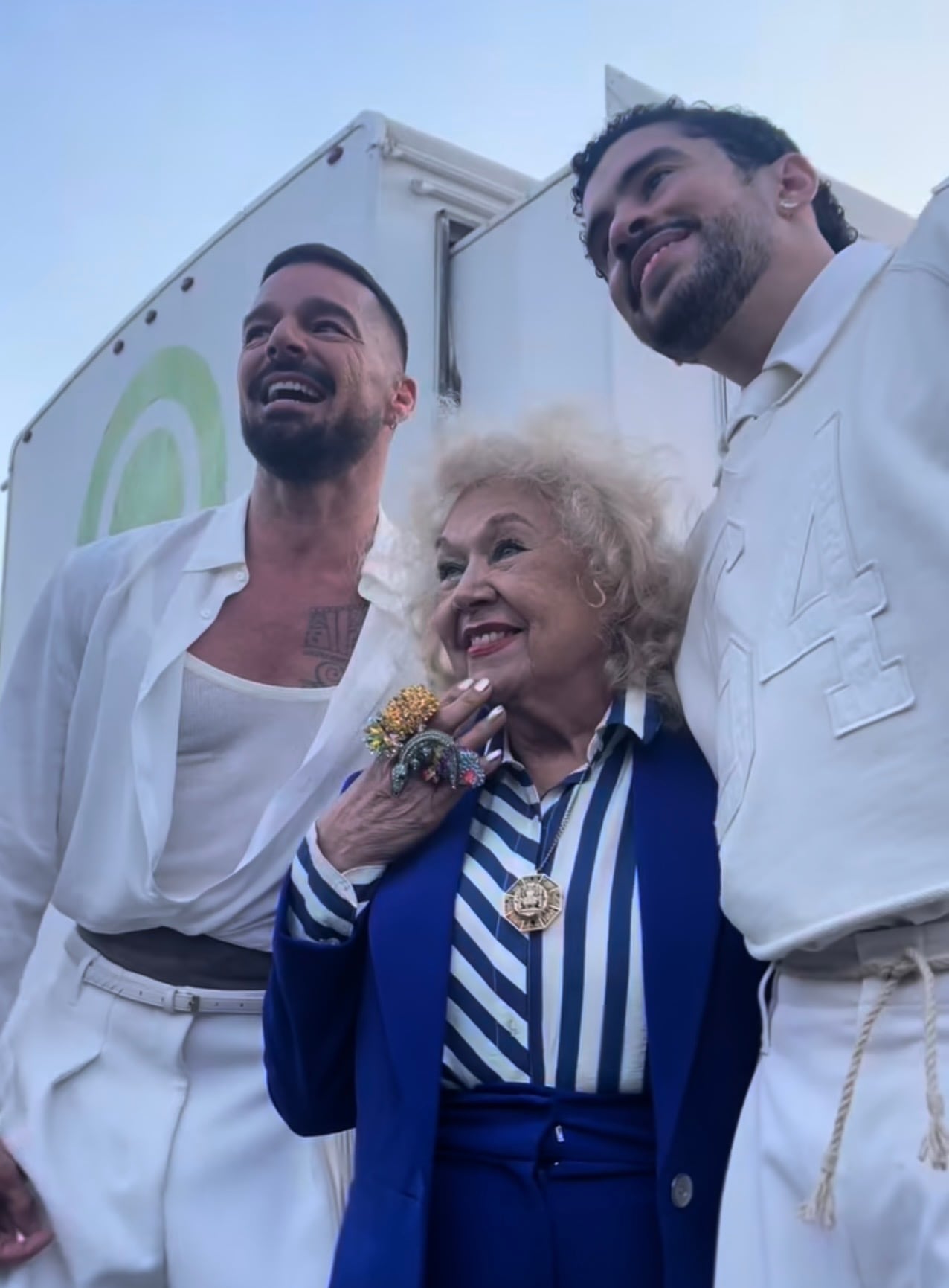 a woman in a blue jacket and pants with big, bold rings poses alongside two men in white