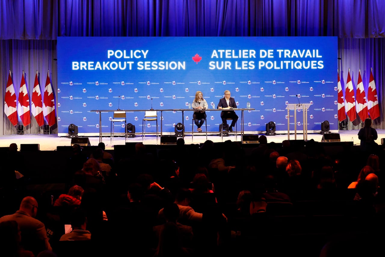Two people sit on a large stage flanked by Canadian flags. A blue backdrop reads 'Breakout policy session'