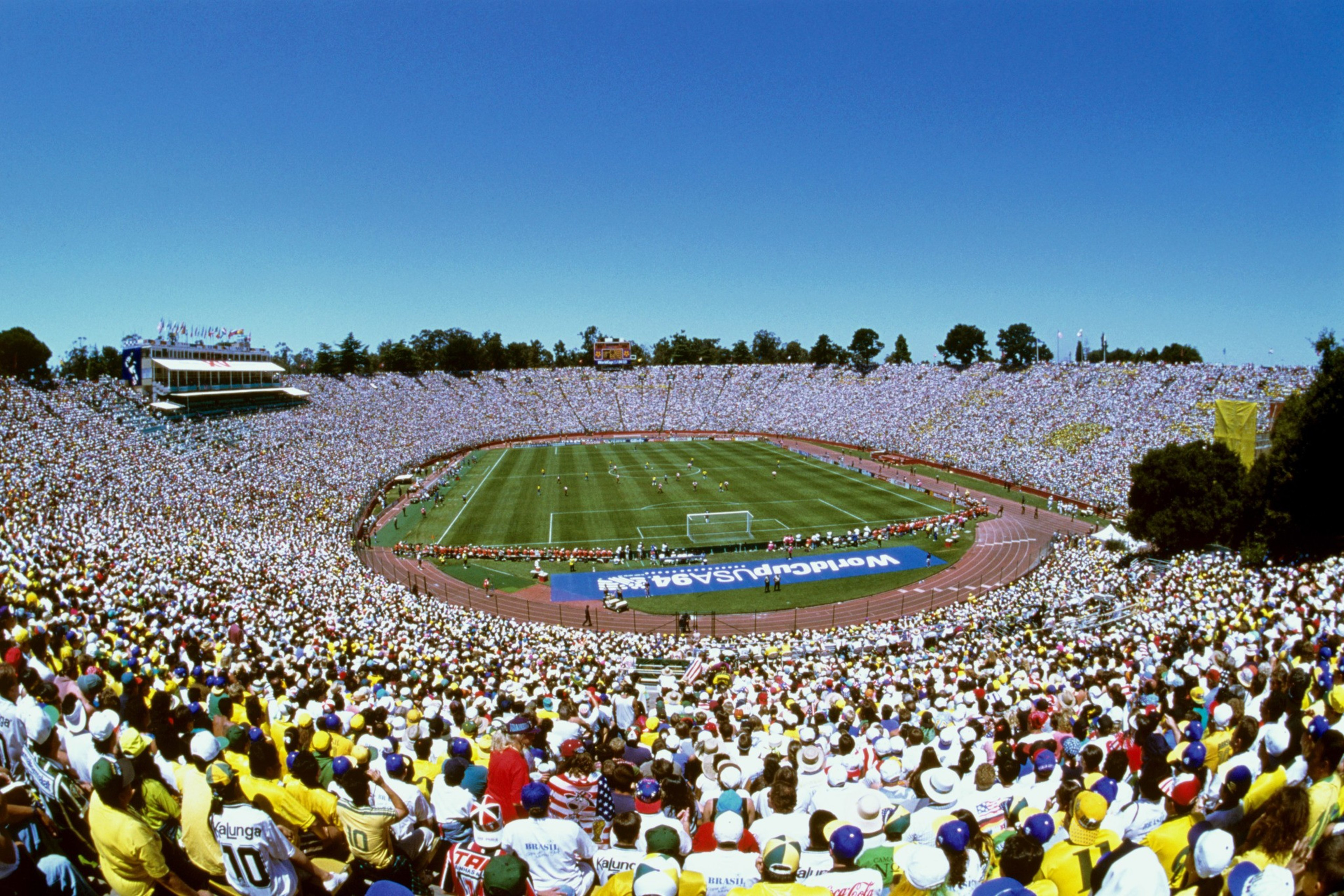 A packed stadium under a clear blue sky hosts a soccer match with thousands of fans wearing colorful shirts and hats in the stands.