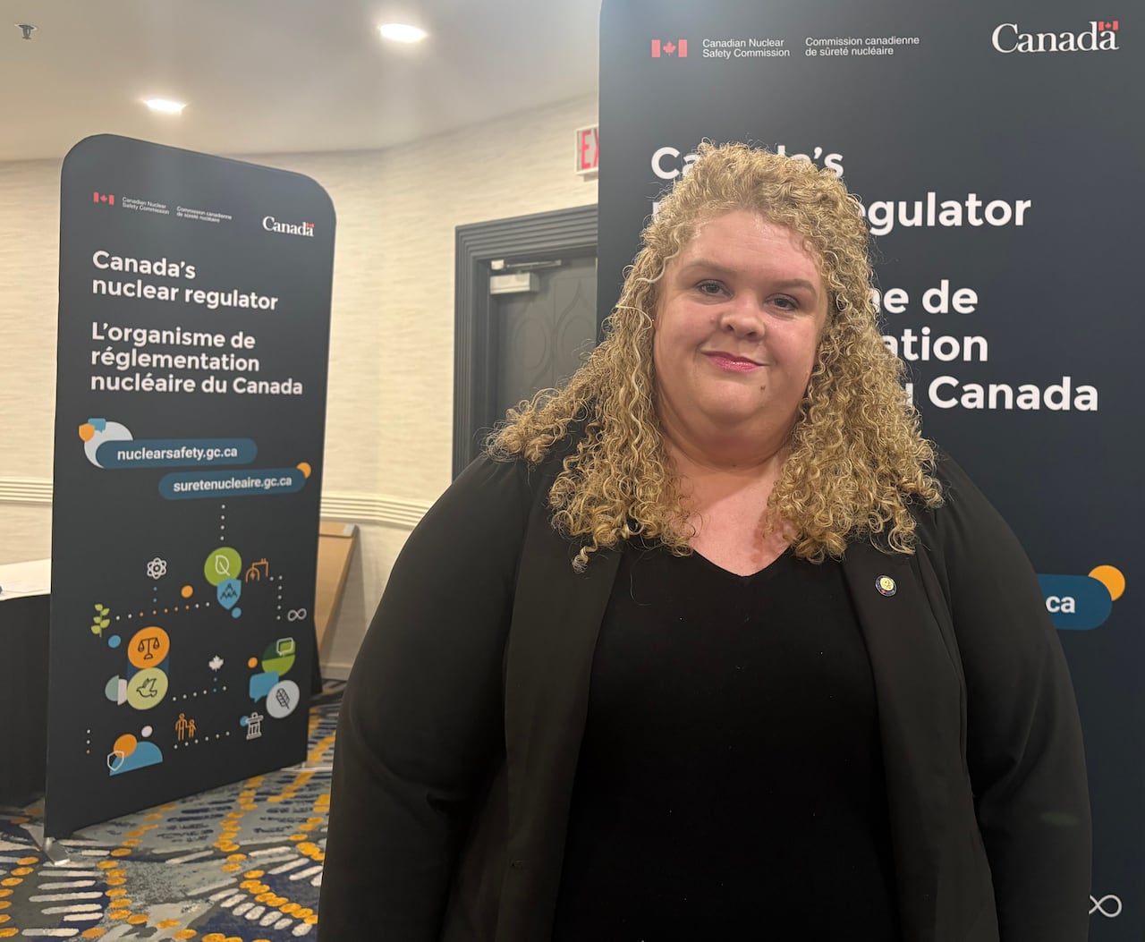 A woman in black clothing with blonde hair stands in front of a federal government sign.