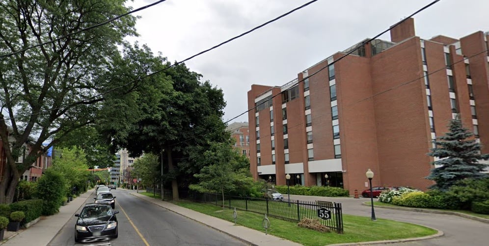 A city street shows residential homes and foliage on one side and a large, brick long-term care home on the other. Two cars are driving down the road toward the camera. It's an overcast spring day