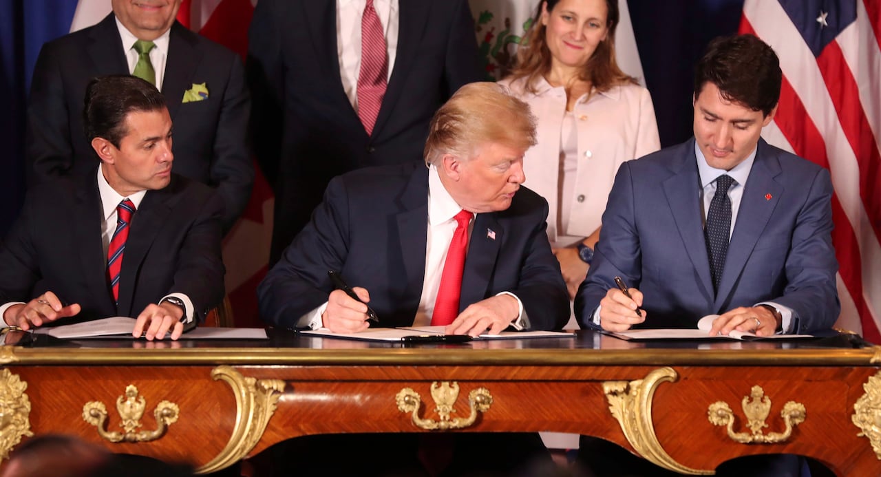 Donald Trump looks toward the paper in front of Justin Trudeau as each holds a pen while seated at a table. 