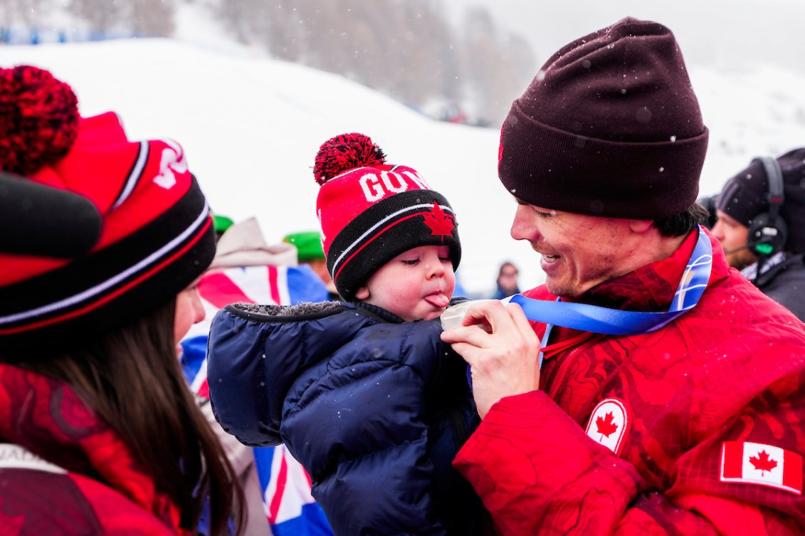 Mikaël Kingsbury of Team Canada celebrates with his young son Henrik after winning silver in freestyle ski moguls at the Milano Cortina 2026 Olympic Winter Games, smiling together in the finish area with snowy slopes in the background.