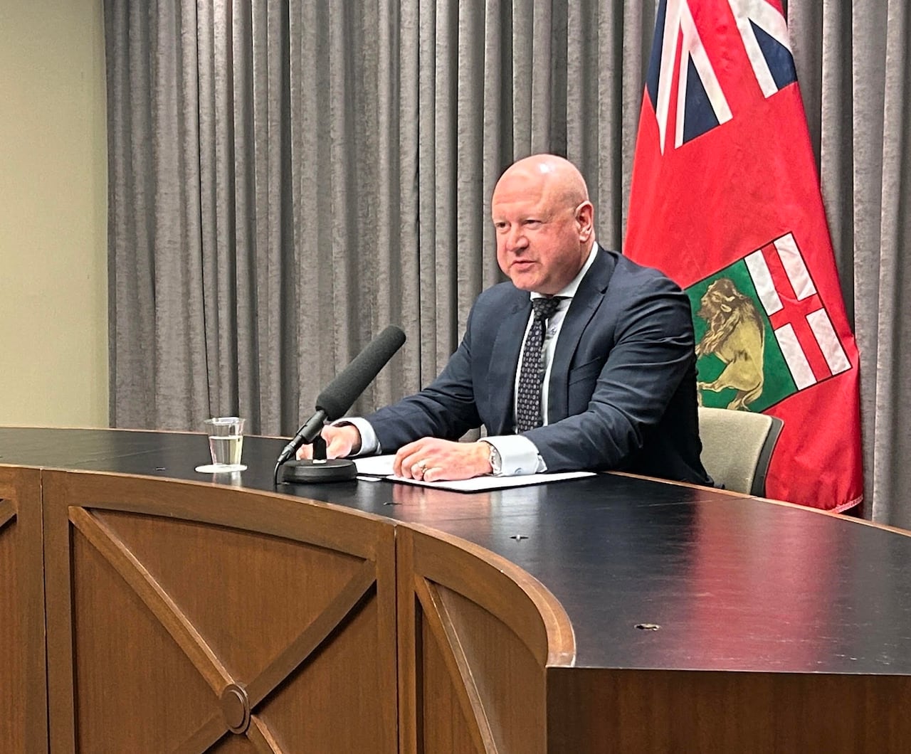 A bald man sits at a desk speaking into a microphone with the flag of Manitoba in the background.