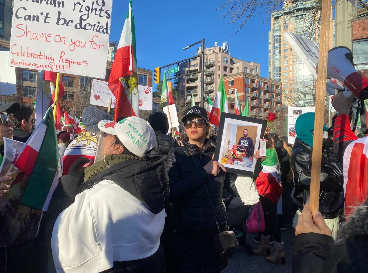 A woman wearing a pair of sunglasses, a cap and a black jacket holds a framed photo of a man as dozens surround her during a demonstration. 