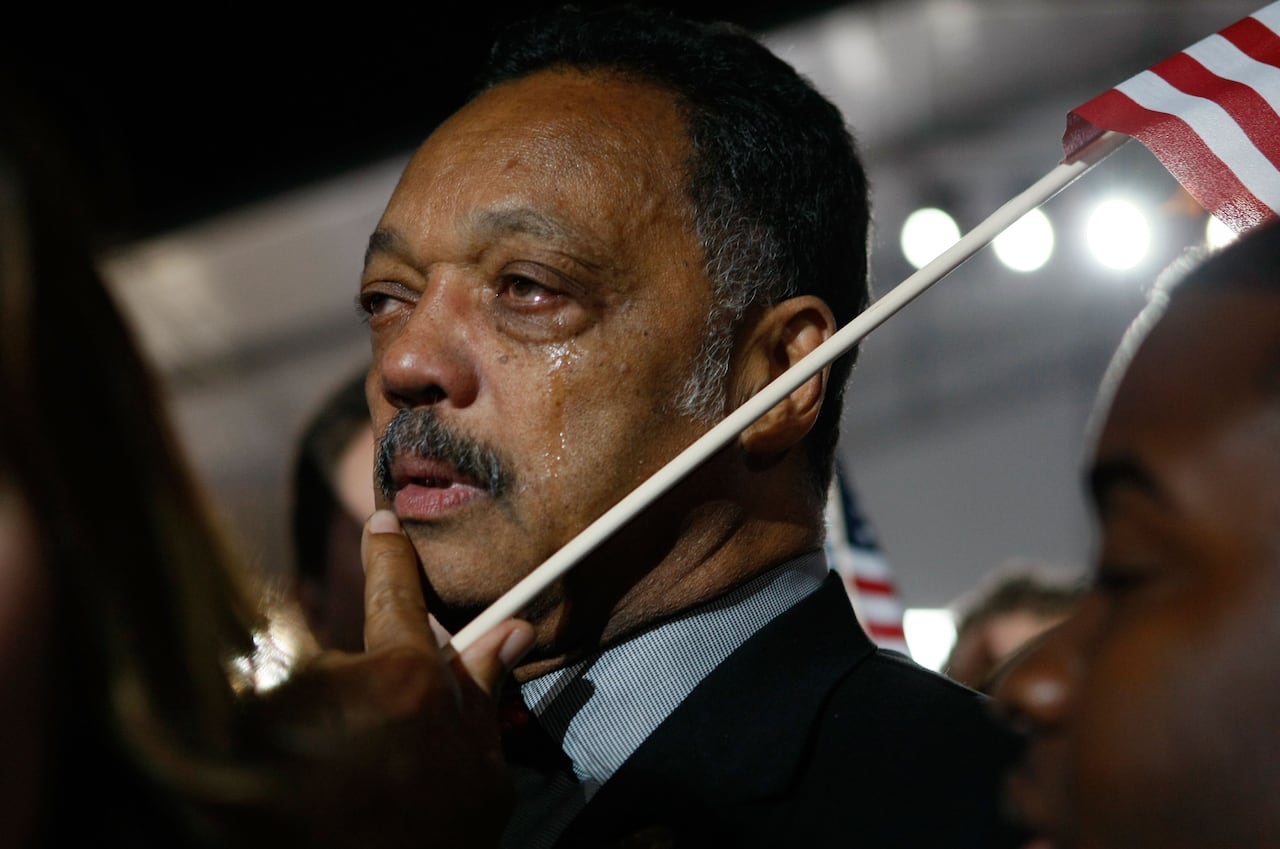A dark-complected man with a mustache has a tear coming out of his left eye while holding an American flag.