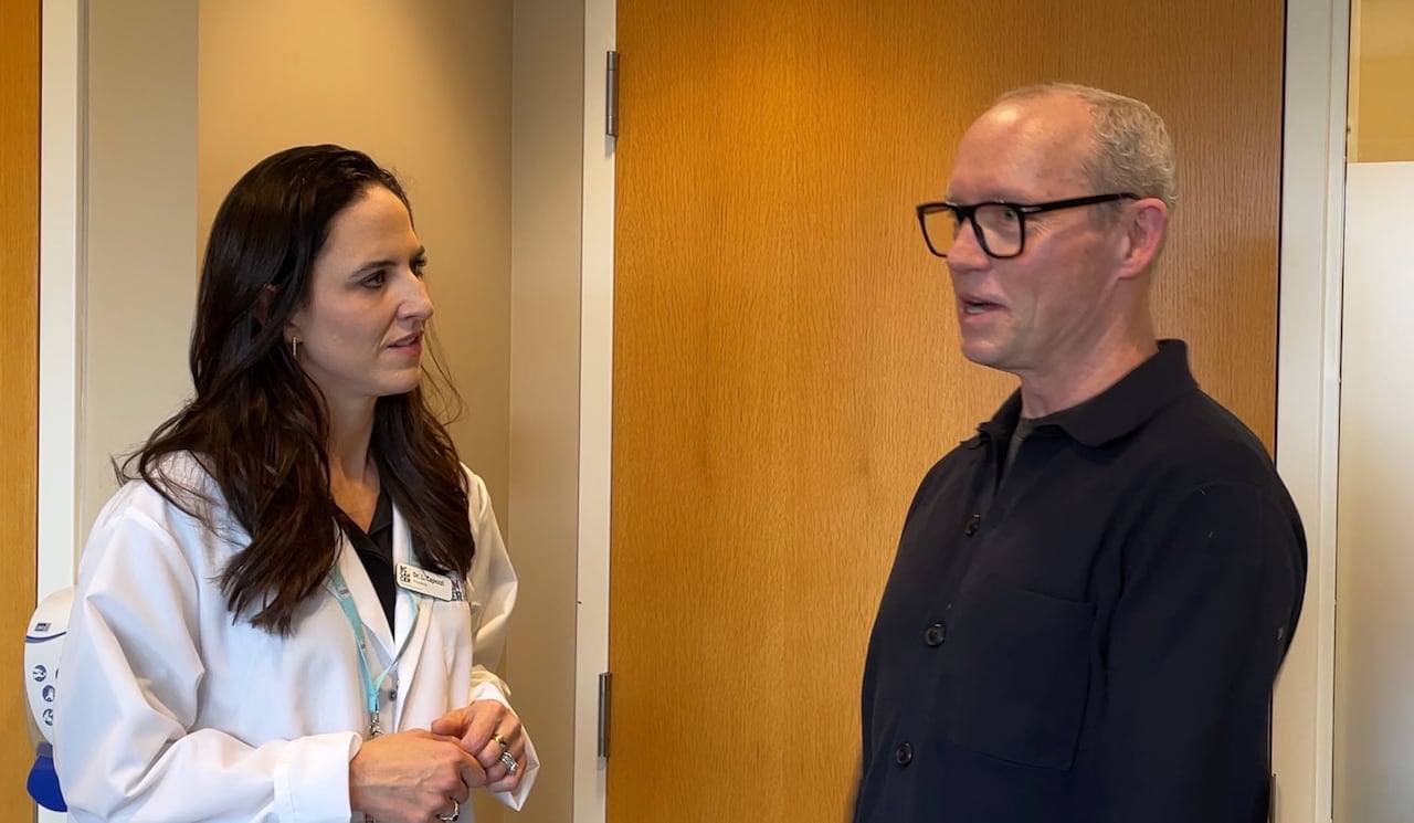 A woman in a lab coat speaks to a man in a black top.