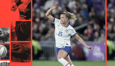A female soccer player in a white USA jersey with number 15 is mid-stride on a grassy field, raising her right arm as she moves forward.