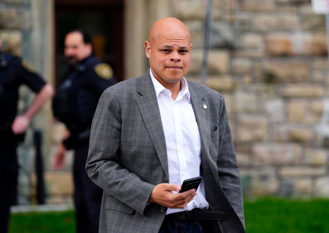 a man in a grey blazer and white shirt holds a phone as he walks outside.