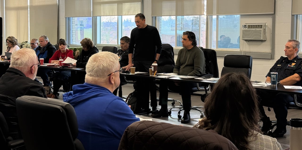 Rows of people sit facing each other behind long tables in a large room. One man is standing up to speak.