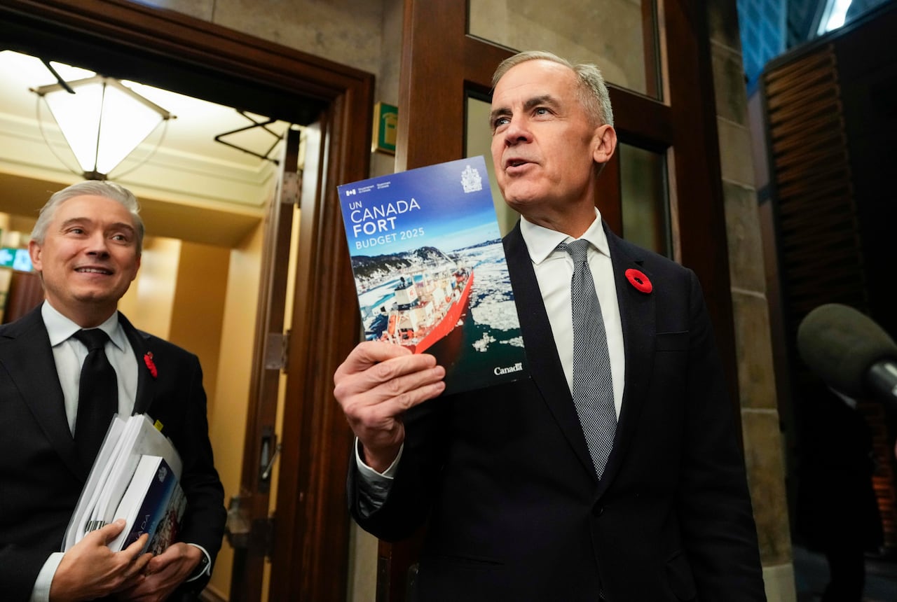 A man in a suit holds up a book that says "Canada Fort" while another man watches on.