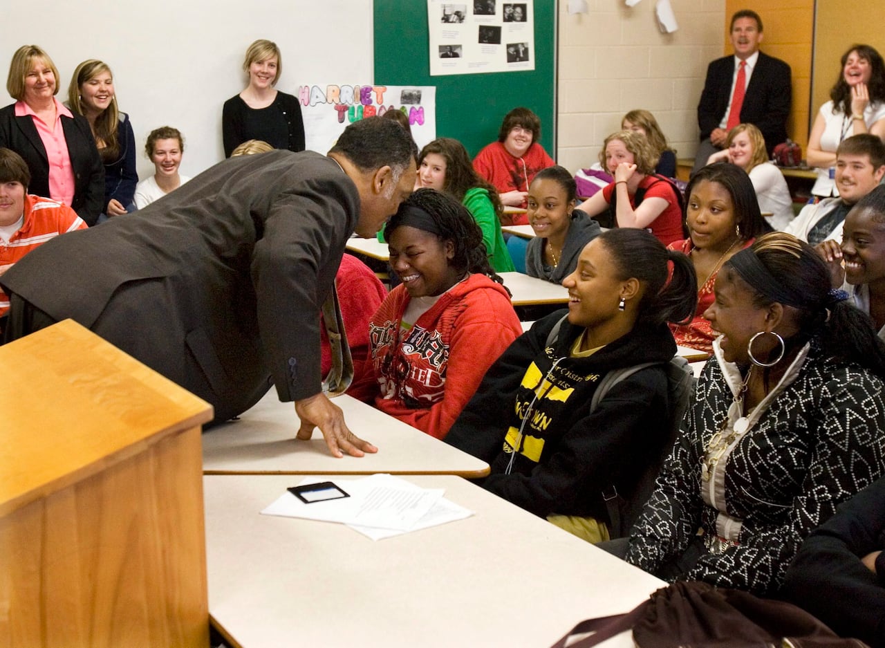 Man stands over a desk and speaks to a girl in a red sweater who is smiling