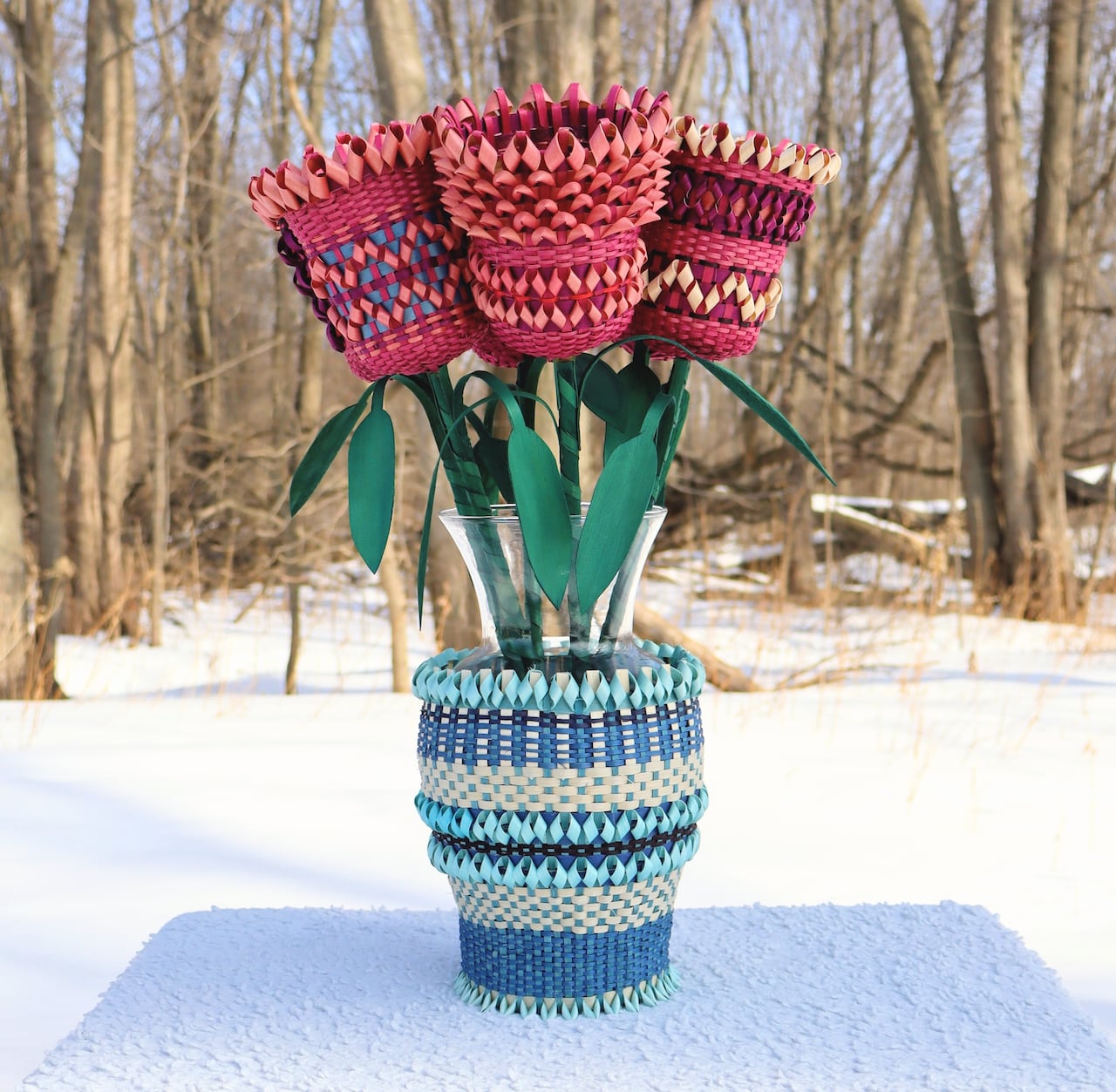 Ann intricately woven blue basket in the shape of a vase, with woven red flowers coming out of the top, sitting in the snow with a forest in the background