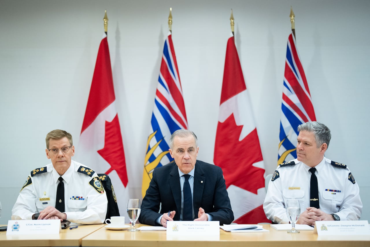 Three men sit at a boardroom table facing forward. The one in the middle wears a dark suit. The two on either side wear white police shirts and black ties. Flags appear in the background.
