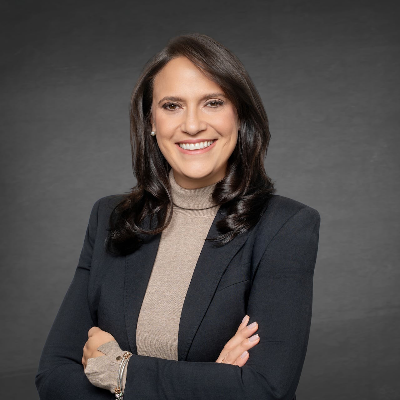 A woman in a blazer and turtleneck stands smiling.