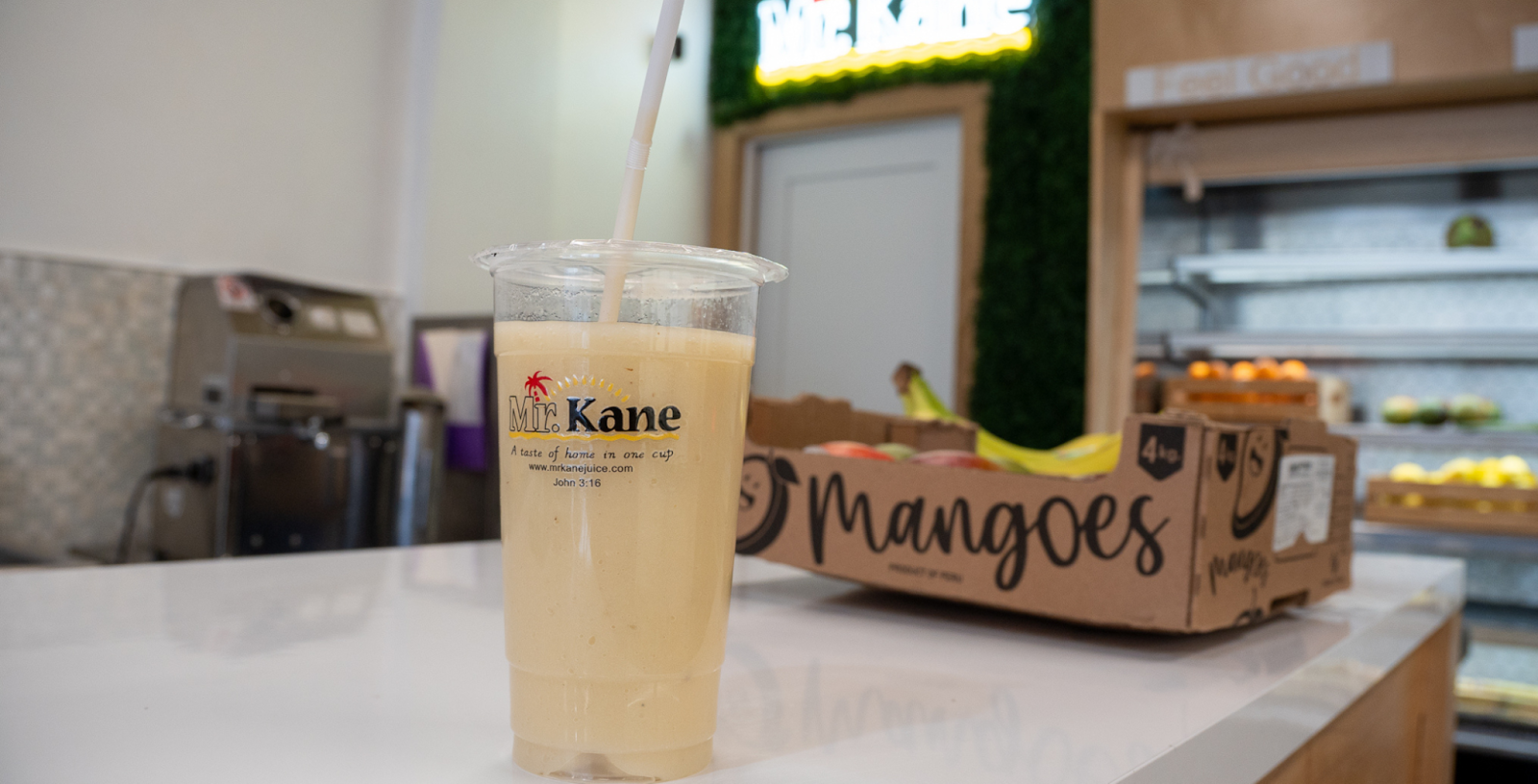 A refreshing mango smoothie in a clear plastic cup with a straw, placed on a white countertop at NOW Toronto, with a box of mangoes and a grocery store background.