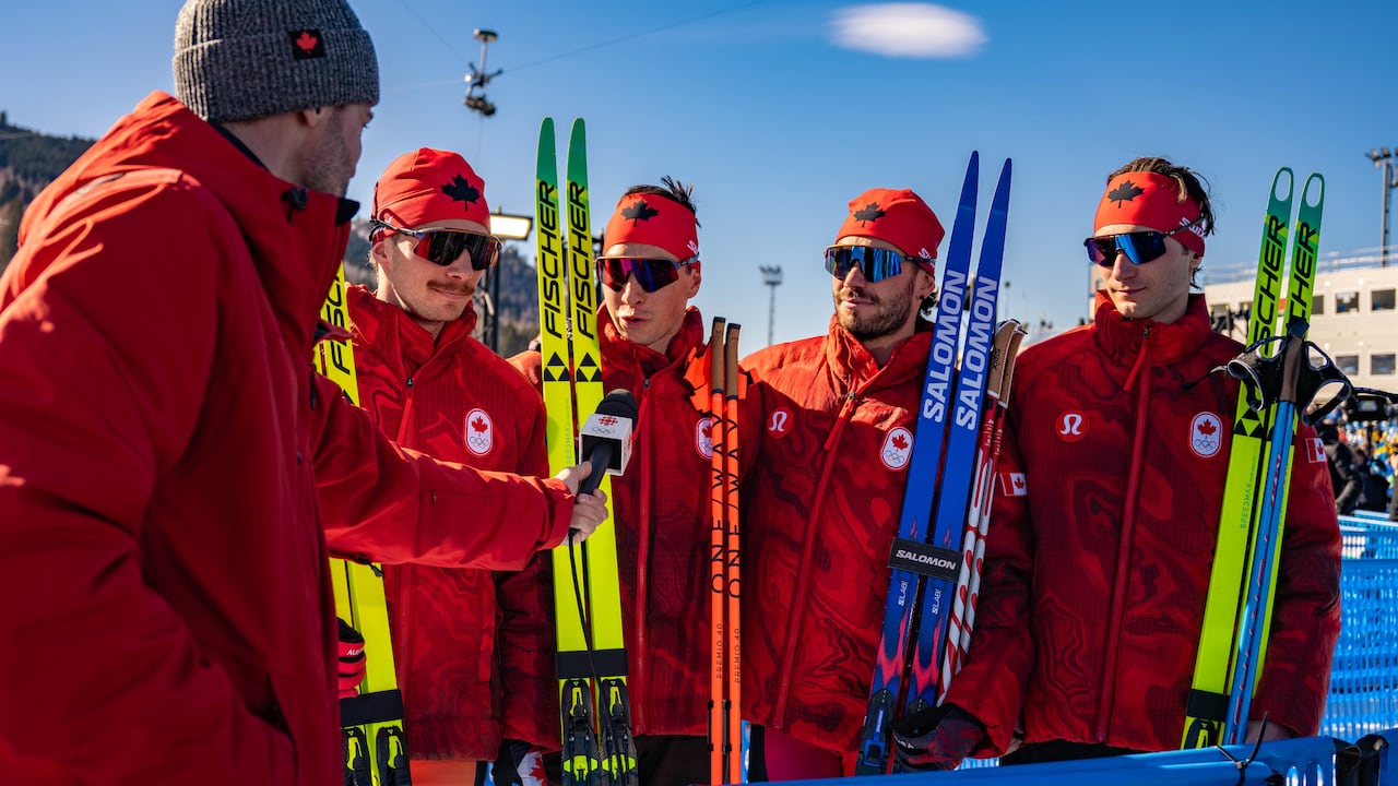 Four men in red outfits holding skis while being interviewed.