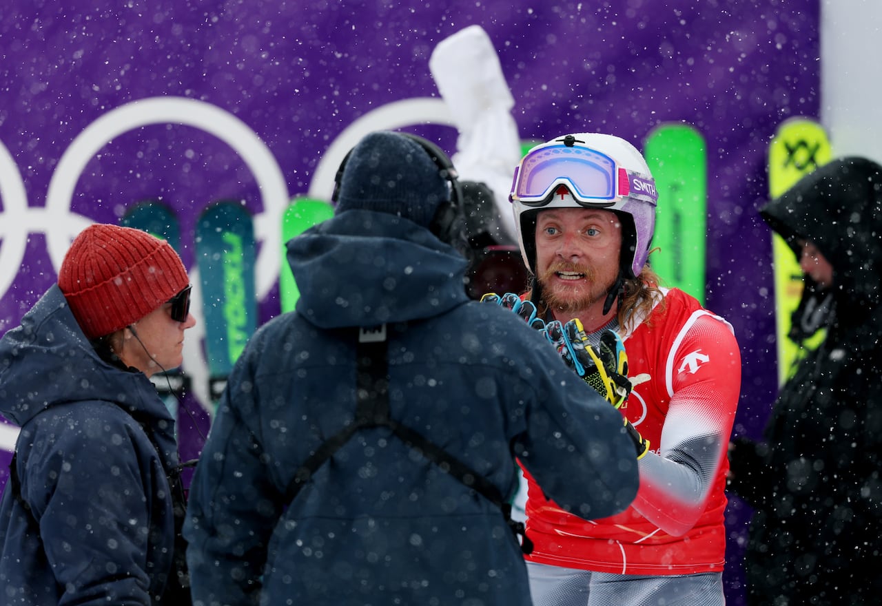A men's ski cross racer raises his hands as a group of officials crowd around him.