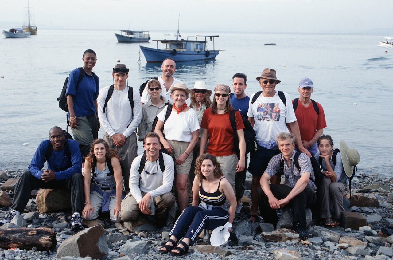 A group of 16 people pose for a picture on a beach.