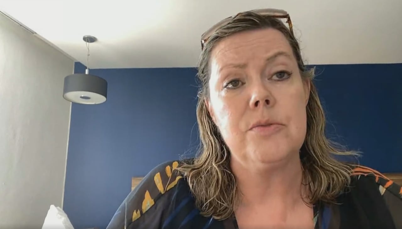 A woman with blonde hair, wearing a black and yellow top sits in a hotel room with a blue accent wall and a lamp.