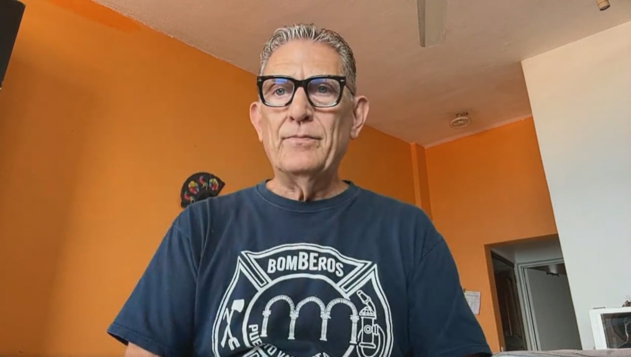 A man with grey hair and black glasses looks forward into a webcam. He is wearing a navy blue t-shirt. The room he sits in has bright orange walls.
