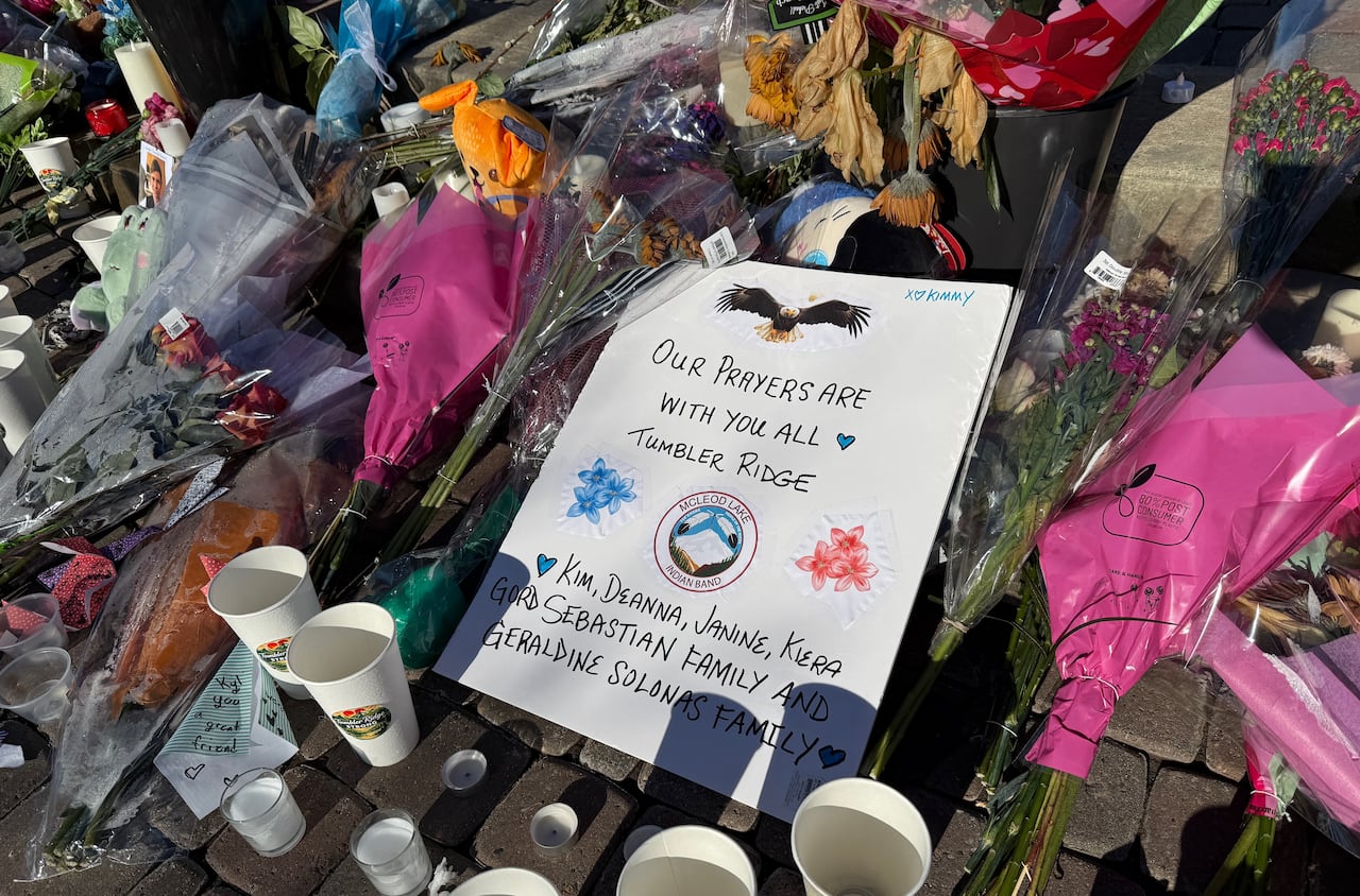 A laminated note lies on a pile of flowers, reading 'Our prayers are with you all.'