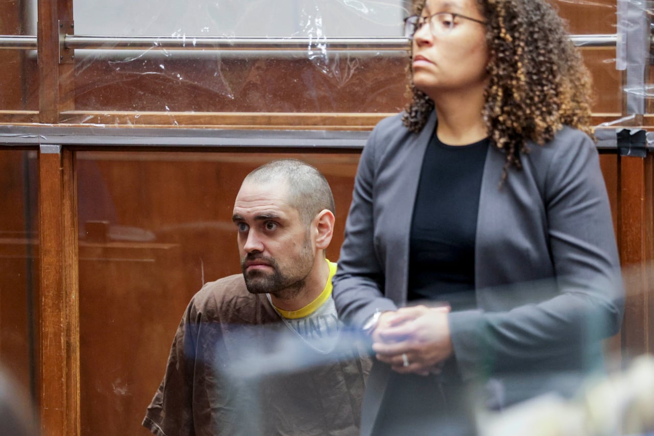 A man in a brown prison outfit sits behind a pane of glass. A woman stands in front of the glass, facing away.
