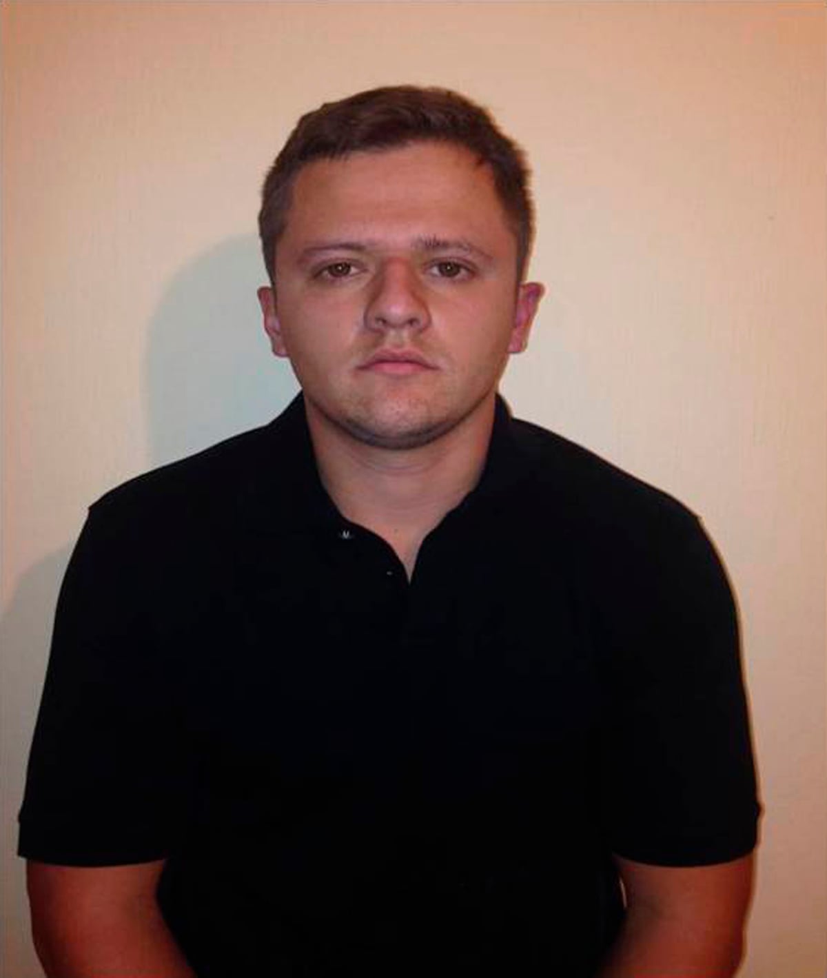 A cleanshaven young man is shown standing while wearing a dark shirt in what appears to be a booking photo, or mugshot.