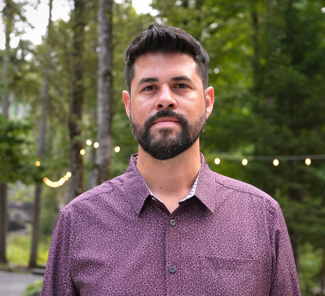 Headshot of a man standing outside, wearing a purple blouse/top. Lights hang in background in the trees.