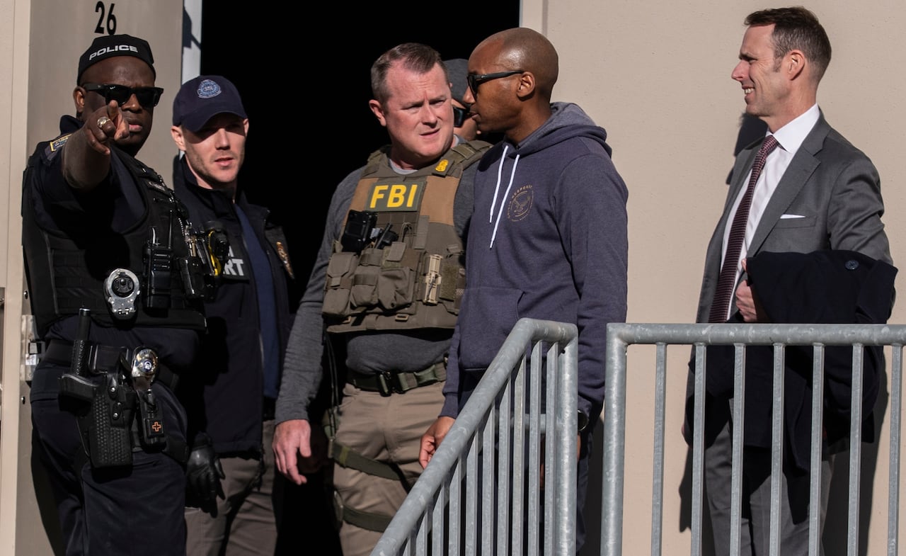 Five men stand outside a building. Three are wearing police uniforms, one a suit and one a grey hooded sweatshirt. 