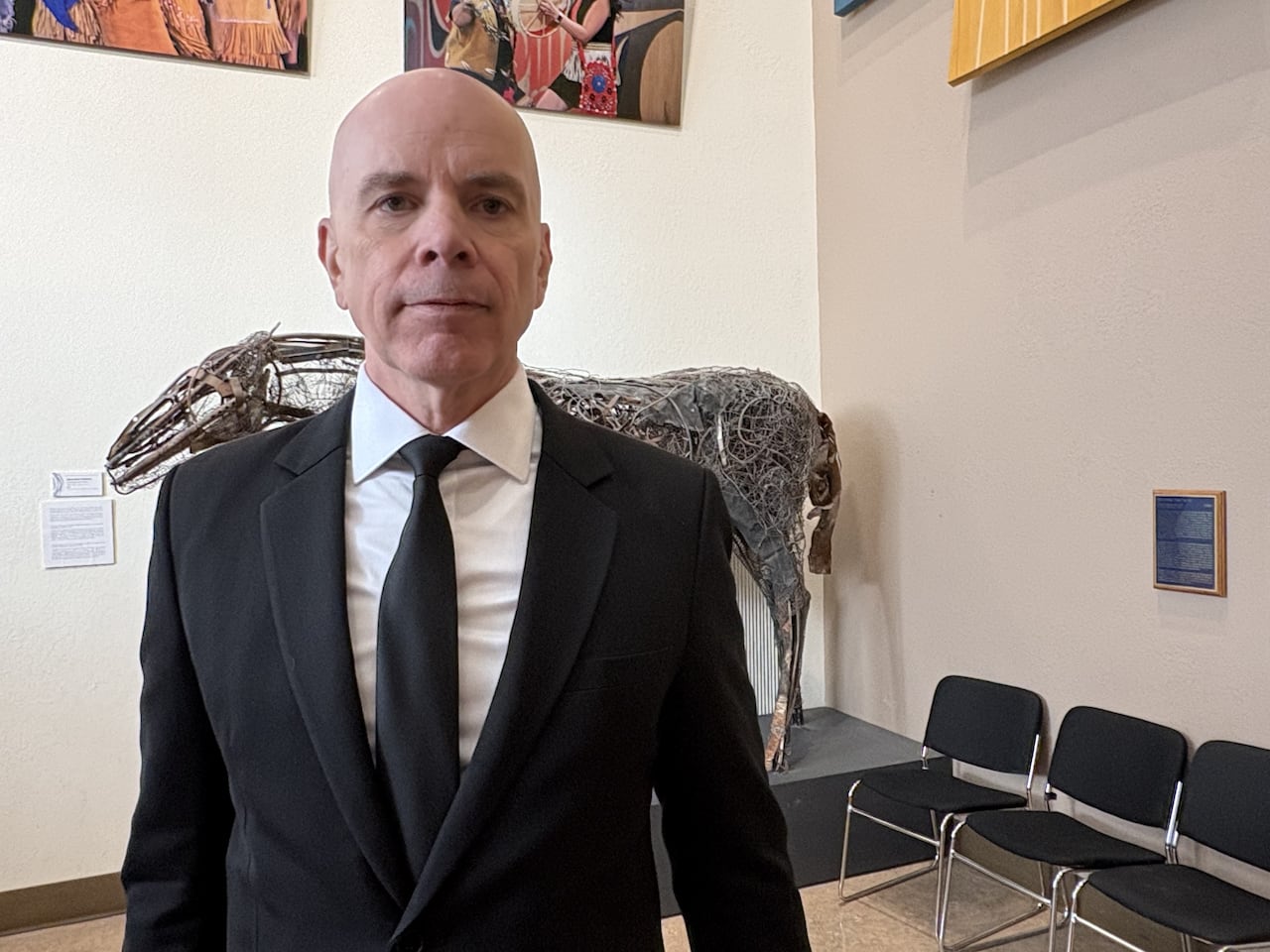 A bald man in a black suit faces the camera.