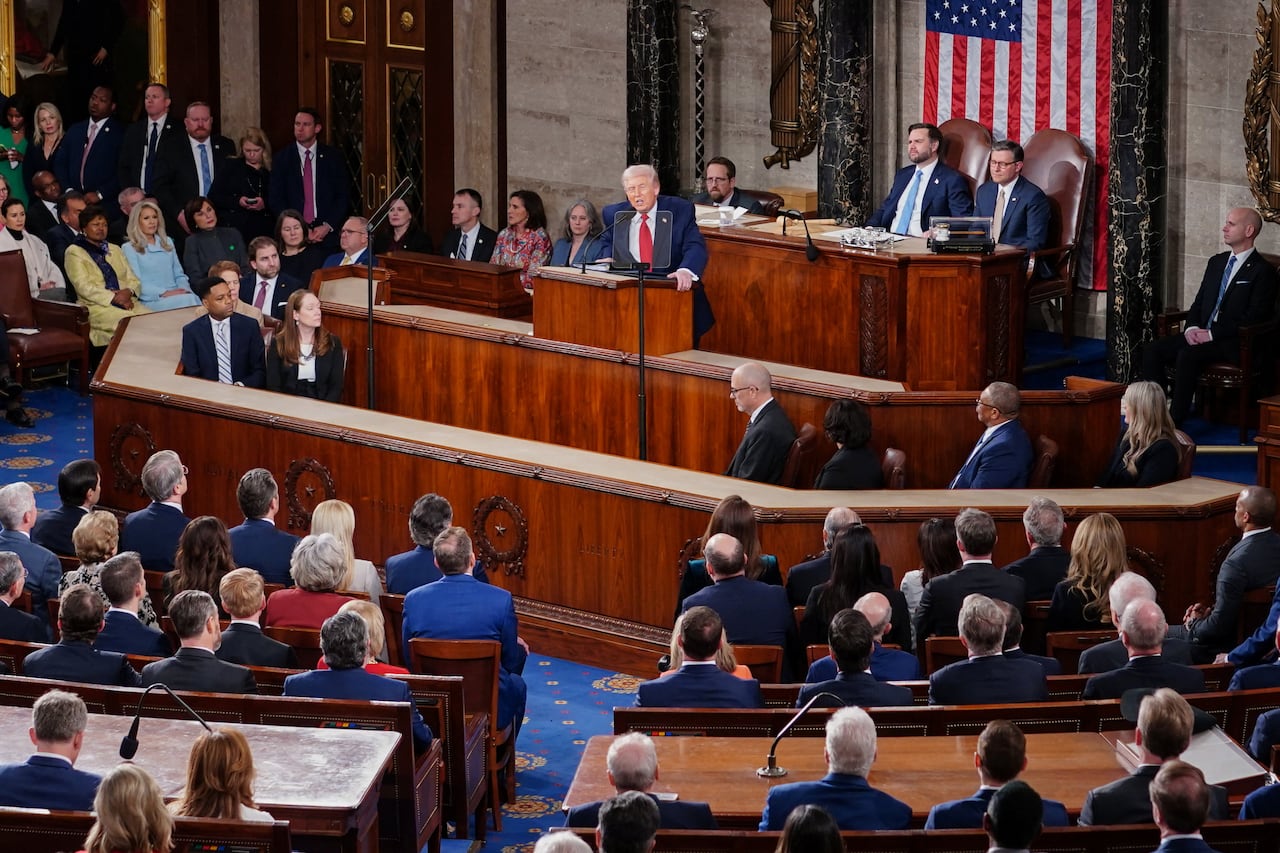 A group of people in suits sit in rows while they watch a man at a lectern give a speech