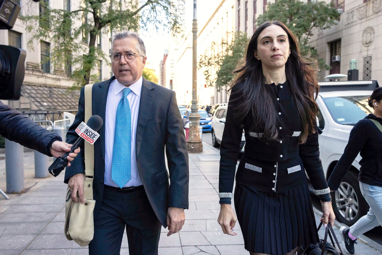 Man in navy suit and blue tie stands left of woman in black blazer and skirt, outside of a mahattan courthouse