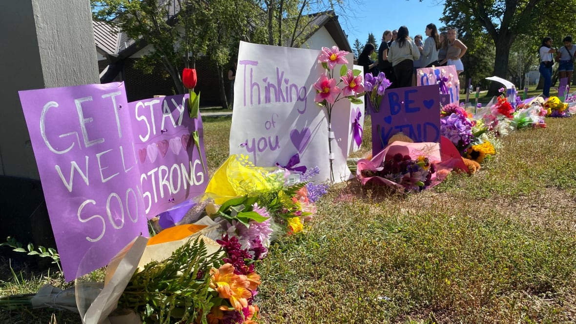 Purple and white signs saying 'Get Well Soon' and 'Stay strong' sit alongside colourful bouquets of flowers in a school yard as students mill around.